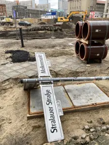 Buntes Treiben in Berlin: Märkte. Einige Holzbohlen der mittelalterlichen Straße wurden bereits herausgerissen, um entsorgt zu werden. Sie liegen am Rand der Ausgrabungsgrube.