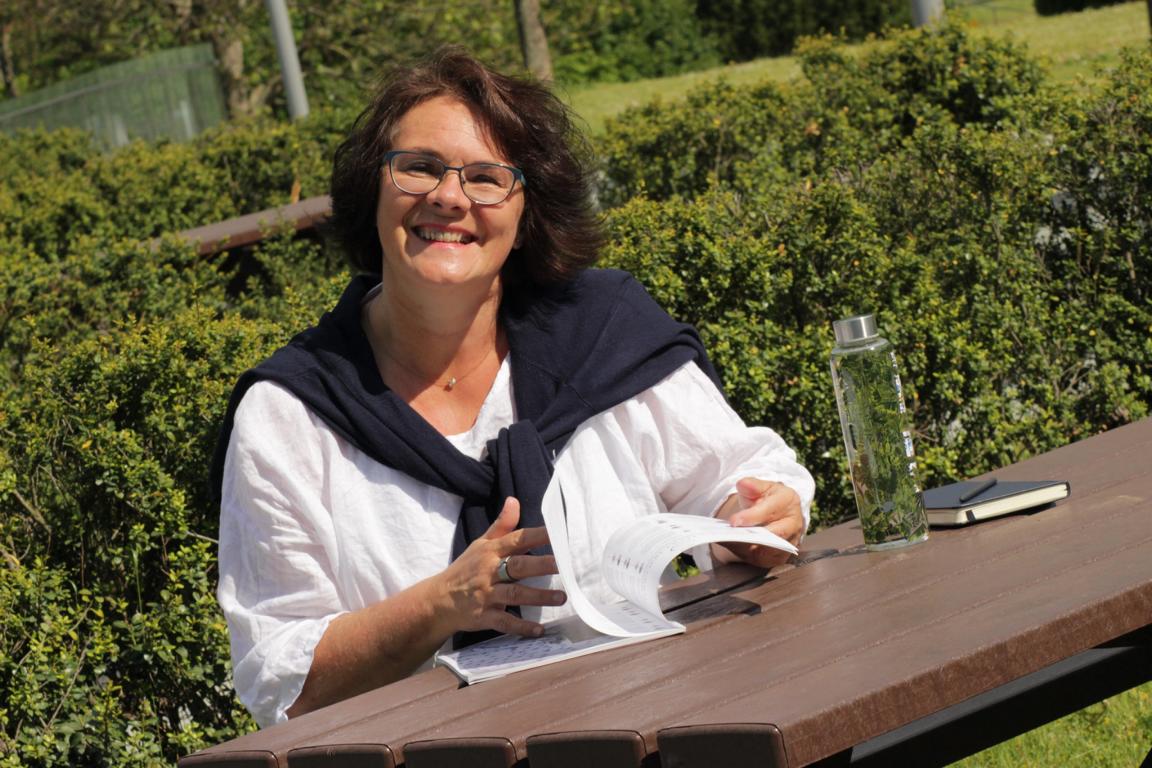 Nicole Isermann schreibt Texte mit Herz und Haltung. Auf dem Bild sitzt sie am Tischdie Zeitung lesend. Das Bild ist bei einem Ausflug entstanden. Es geht sommerlich zu.