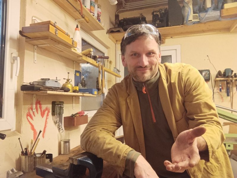 Christian Abt in seiner Werkstatt. Im Vordergrund seine Hand, die die Handhabung symbolisiert, weil er für ge-hand-h-abt steht. Er ist in seinem beigen Jackett leger gekleidet, ein Mann in den 40igern.