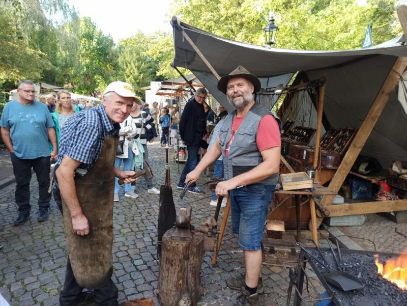 Falk Laxander ist Kunst- und Silberschmied. Bei KUNST trifft WEIN zeigt er Besuchern, wie das Schmieden geht. Auf dem Bild sehen wir ihn mit einem erwachsenen Besucher, mit dem er zusammen an einem Werkstück arbeitet.