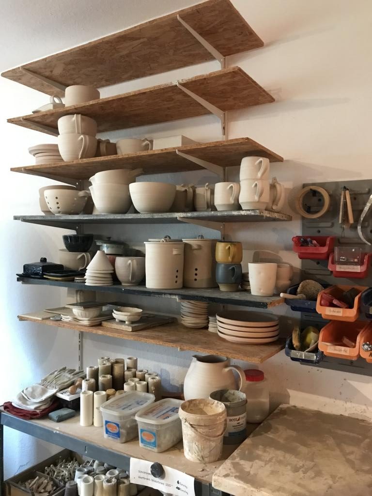 Regalbretter an der Wand. Darauf stehen allerlei Vasen, Römertöpfe, also eher größere Werkstücke, die trocknen sollen.