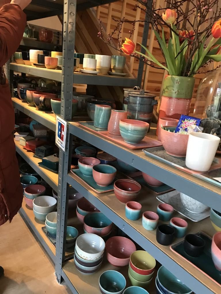 Ein Regal voller farbenfroher Vasen, Teller, Tassen mit und ohne Henkel stehen bereit gekauft zu werden. Es gibt frische Tulpen in der Vase.