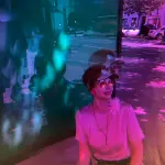 Betty setzt vor einem Schaukasten mit grün gestrichener Fläche. Sie hat die Wheels ihres Rollstuhls pink eingekleidet und brilliert mit geschmackvoller Kleidung.