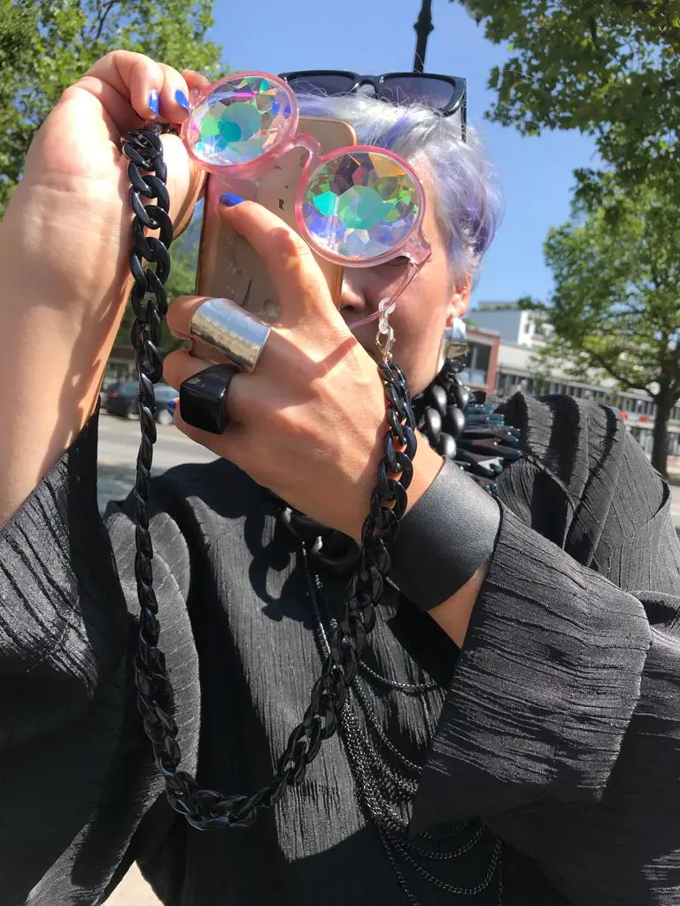 Daria Fürst fotografiert durch ihre Prismabrille. Sie ist durchgängig schwarz gekleidet. Halskette und Umhängekette, an der die Brille hängt, erinnern an ein Vorhängeschloss an der Wohnungstür.