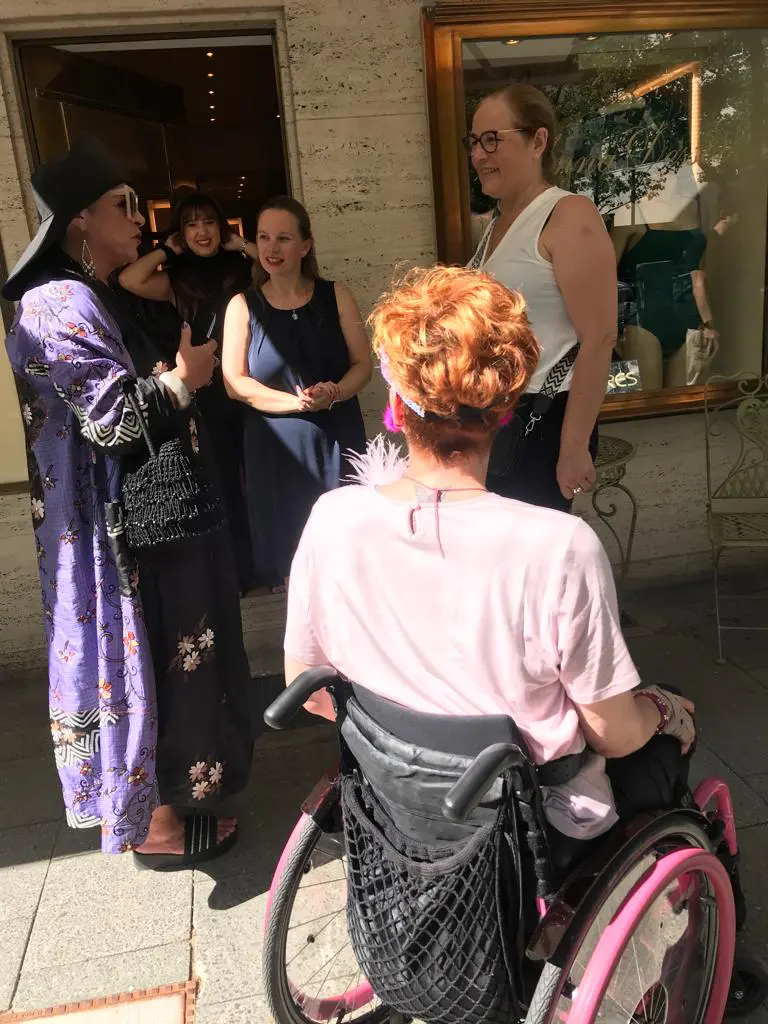 Walk of Glam trifft auf Begeisterung der ortsansässigen Geschäftsleute am Ku'damm: im Vordergrund Betty im Rollstuhl, links Stella Ahangi und dahinter die frohgelaunten Angestellten und Kunden einer Modeboutique.