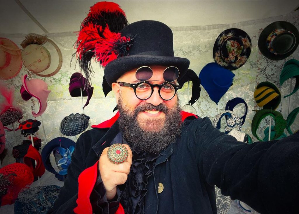 Gianni Klein im selbstbewussten Vintage-Look vor seinem Marktstand voller Hutkunst – ein Selfie zwischen Stil, Spiel und Statement.