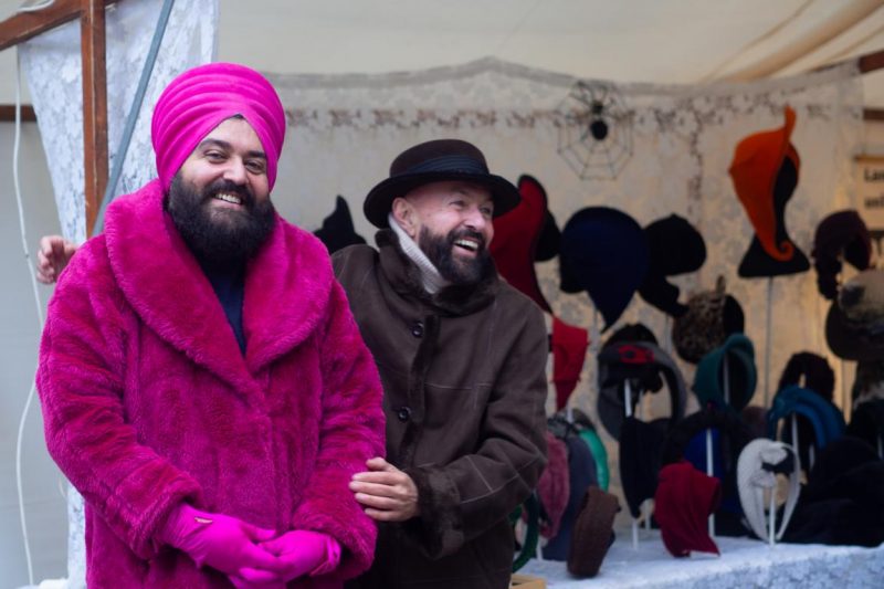 Gianni in pinkem Turban und Pelz neben Carlos – ein Duo, das Mode zur Begegnung macht und Hüte zur Haltung.
