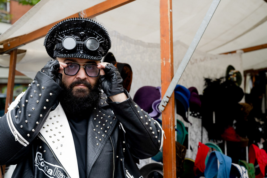 Mann mit Spikes und Sonnenbrille vor seinem Hutstand; seine Jacke trägt „Loyalty“, der Hut wirkt wie ein rebellisches Statement.