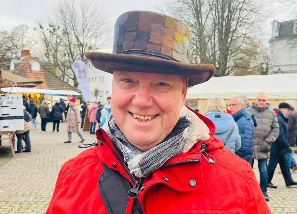 Ein Mann mit Patchworkhut und rotem Mantel lächelt auf dem WEInachtsmarkt – zufrieden mit seinem neuen Look.